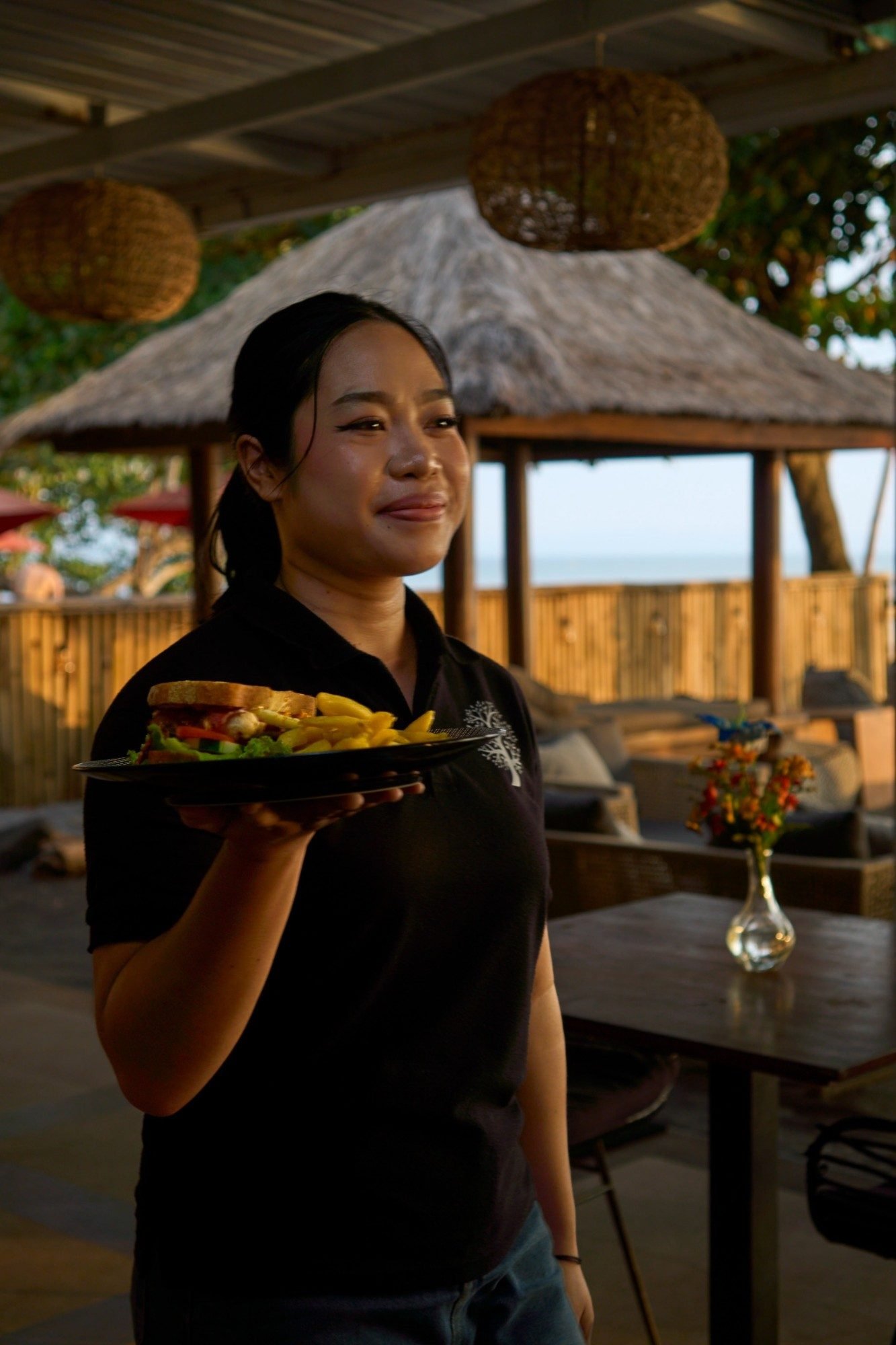 A JOJE Beach Bar & Resto team member serving a sandwich and fries at sunset
